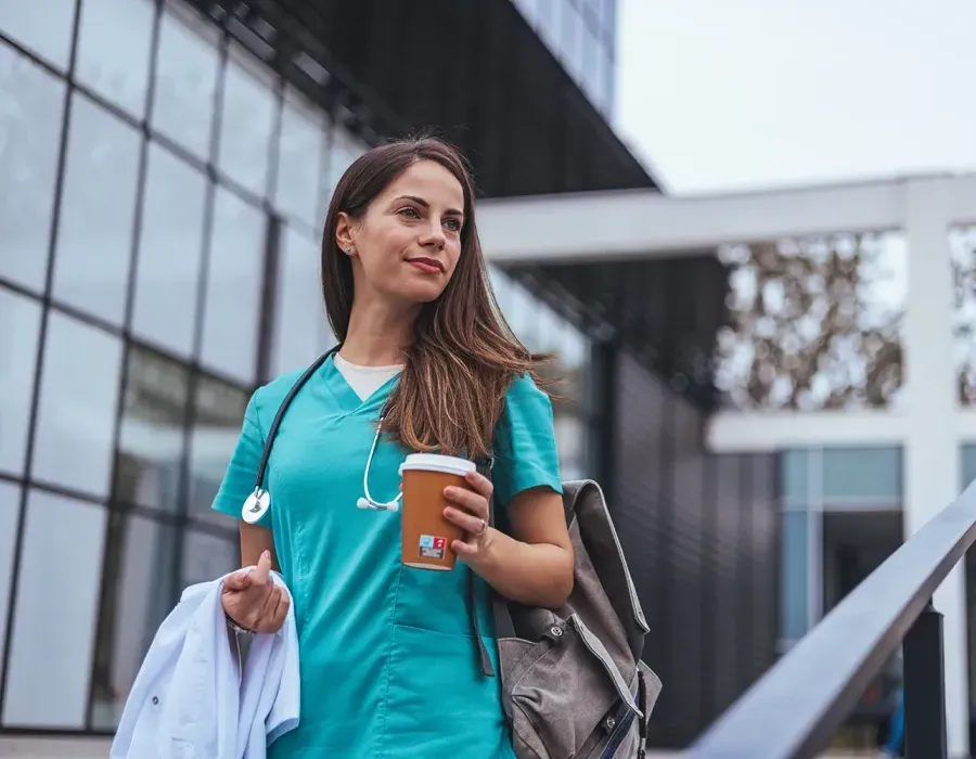 Woman travel nurse wearing green scrubs and backpack bringing coffee to work